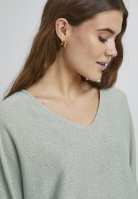 Hellgrüner gestrickter Pullover mit V-Ausschnitt, der eine weiche Textur und eine lässige Passform bietet. Auch goldene Creolen sind sichtbar.