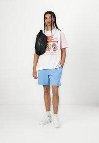 Sort skuldertaske, lyserød T-shirt med grafik, der viser tegnefilmsfigurer, blå shorts, hvide sneakers, sat mod en simpel hvid baggrund.