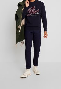 Maglione blu navy con grafica rossa e bianca "Pepe Jeans", pantaloni blu navy scuri, sneakers alte color crema, e un parka verde tenuto su un braccio.