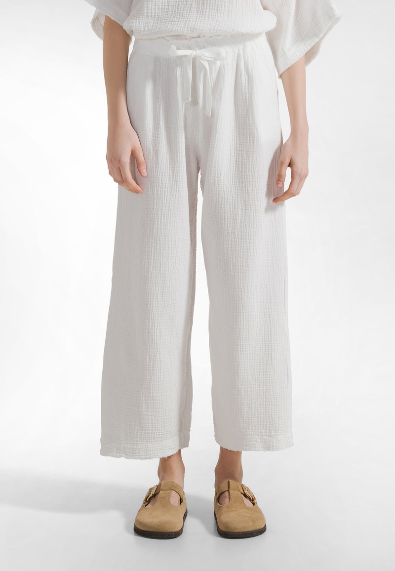 Henkilö, jolla on päällään väljät valkoiset, tekstuurilliset housut, joissa on kiristysnyöri vyötäröllä, ja jaloissa vaaleanruskeat mokkaiset slip-on-kengät valkoista taustaa vasten.