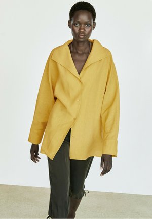 Mørkhudet kvinde iført en oversized sennepsgul skjorte med knapper og afklippede sorte bukser, går mod en ensfarvet lys baggrund.