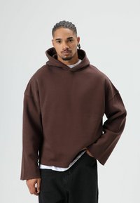 Sweat à capuche oversize marron avec une texture lisse et des détails à bord brut, superposé à un t-shirt blanc, associé à un pantalon foncé.