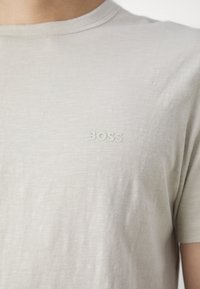 T-shirt beige clair en tissu doux, avec un col rond et un logo "BOSS" discret sur la poitrine gauche. Texture lisse, design décontracté.