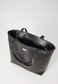 Sac fourre-tout en cuir noir avec deux poignées, fermeture éclair et poche intérieure. Présente une petite étiquette de logo en rouge, blanc et bleu.
