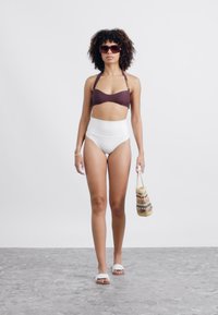 Brauner Bikini-Oberteil kombiniert mit hochgeschnittenen weißen Badehosen. Modell trägt runde Sonnenbrillen und hält eine gemusterte Tasche. Weißer Hintergrund.