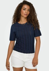 Navy blauwe gebreide top met korte mouwen, voorzien van een geribbelde structuur, scalloped halslijn en zoom, gecombineerd met witte shorts.