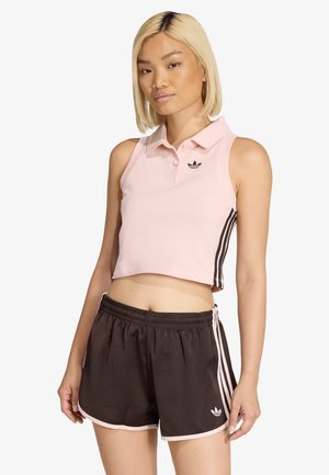 Kobieta o blond włosach, ubrana w różowy krótki top polo bez rękawów i ciemnobrązowe sportowe szorty z różowymi paskami i logo.