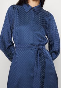 Mörkblå klänning med geometrisk struktur, långa ärmar, krage och ett knytband i midjan. Slätt tyg med subtil lyster.