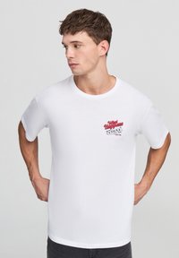 Biała bawełniana koszulka z krótkim rękawem, z okrągłym dekoltem, z czerwonym i czarnym nadrukiem na lewej piersi mówiącym "Co się dzieje w Vegas".