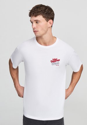 T-shirt in cotone bianco con una stampa grafica rossa e nera che recita "Cosa succede a Las Vegas" sulla parte sinistra del petto. Maniche corte, scollo rotondo.