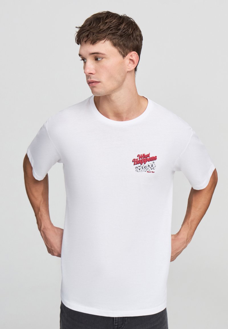 Biała bawełniana koszulka z krótkim rękawem, z okrągłym dekoltem, z czerwonym i czarnym nadrukiem na lewej piersi mówiącym "Co się dzieje w Vegas".