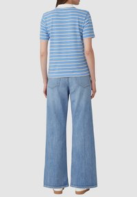 Frau, die ein hellblau gestreiftes Poloshirt und weit geschnittene hellblaue Jeans trägt, steht mit dem Rücken zu einer einfarbigen Hintergrund.