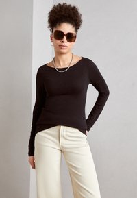 Haut noir à manches longues avec un décolleté off-shoulder allongé, associé à un pantalon large crème et des lunettes de soleil marron surdimensionnées.