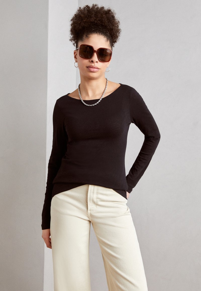 Haut noir à manches longues avec un décolleté off-shoulder allongé, associé à un pantalon large crème et des lunettes de soleil marron surdimensionnées.