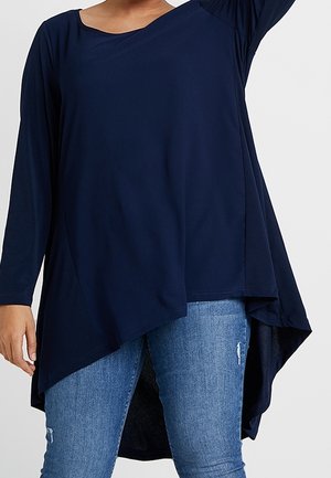 Personne portant un haut tunique asymétrique à manches longues bleu marine avec un jean bleu clair déchiré sur un fond uni.