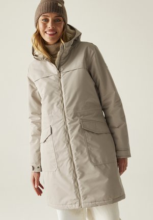 Donna sorridente che indossa un cappotto invernale beige con grandi tasche e un berretto marrone lavorato a maglia, in piedi davanti a uno sfondo chiaro e semplice.