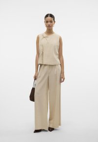 Vrouw met beige mouwloze top en wijde broek, houdt een kleine bruine handtas vast tegen een effen witte achtergrond.