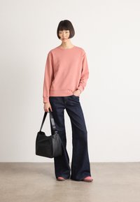 Woman with short dark hair wearing a pink sweatshirt, wide dark blue jeans, pink shoes, and holding a black handbag, standing on a beige floor.