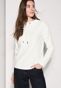 Camiseta blanca de manga larga con rayas texturizadas, que presenta una capucha y acentos de cordón. Combinada con pantalones de mezclilla azul oscuro.