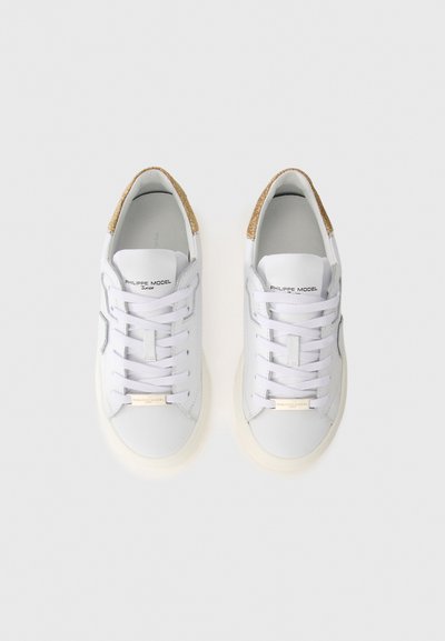 White leather sneakers with a textured gold accent at the heel, featuring six pairs of eyelets, flat white laces, and a subtle logo on the tongue.