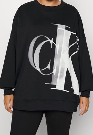 Osoba ubrana w czarną, oversize'ową bluzę z dużymi, nałożonymi na siebie białymi i szarymi literami "CK" na przodzie, w połączeniu z czarnymi spodniami.