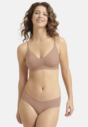 Donna con capelli mossi che indossa un reggiseno nude senza cuciture e biancheria intima abbinata, in piedi contro uno sfondo chiaro semplice, con un leggero sorriso.