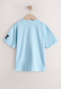 Tricou bleu deschis, cu mânecă scurtă, agățat de un umeraș de lemn pe un perete bej simplu, cu un desen negru pe mâneca stângă.