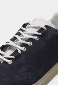 Baskets en daim bleu marine avec lacets blancs passant à travers des œillets métalliques, languette blanche rembourrée et semelle en caoutchouc beige.