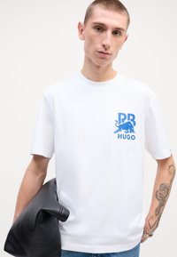T-shirt bianco in cotone con maniche corte presenta un motivo grafico blu nell'area del petto sinistro. Il modello tiene un accessorio in pelle nera.