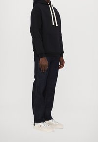 Persona con una sudadera negra con cordones blancos, jeans oscuros y zapatillas blancas de pie contra un fondo blanco liso.