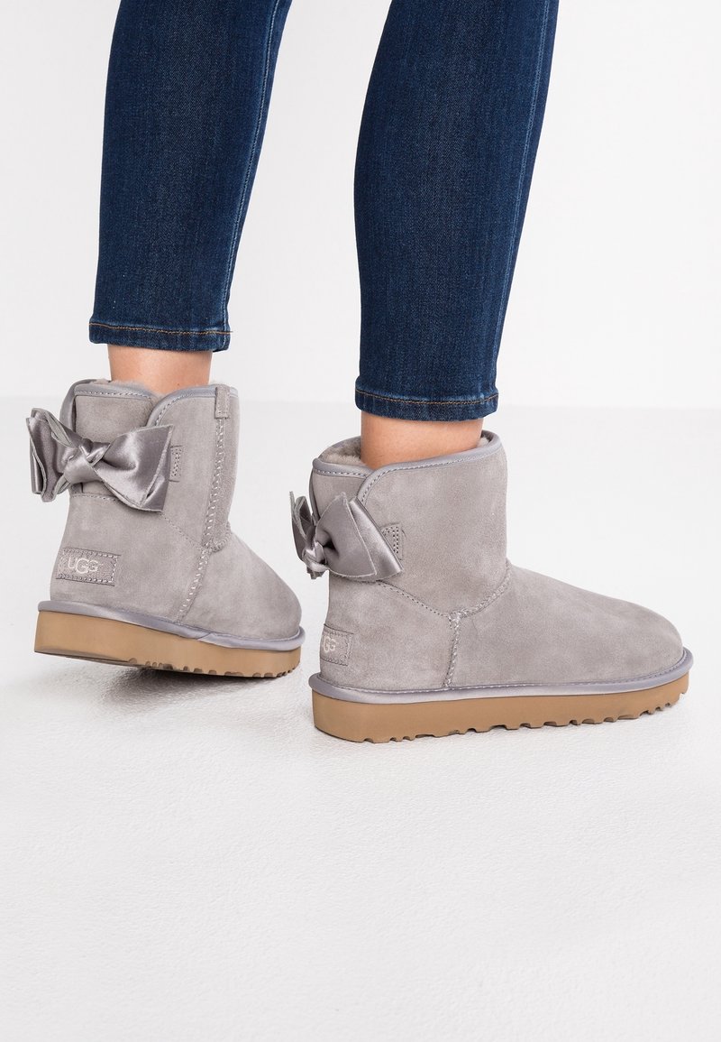 Gray suede ankle boots with a flat sole, featuring a decorative satin bow on the back and a logo patch on the side.