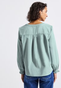 Femme aux cheveux bouclés portant un haut à manches longues vert clair et un jean bleu, vue de dos contre un fond uni.
