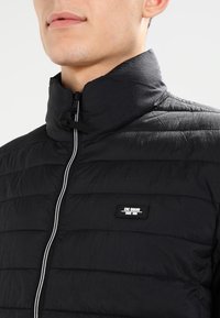 Černá prošívaná bunda s vysokým límcem, předním zipem a značkovým štítkem. Má texturovaný povrch a bílý akcent na zipu.