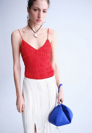 Femme portant un haut rouge à bretelles spaghetti froncé et une jupe blanche, tenant un sac à main souple triangulaire bleu sur un fond uni.