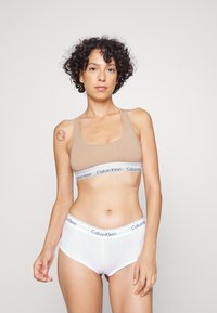 Bralette sportivo nudo con spalline larghe e fascia con logo Calvin Klein bianco; slip hipster bianchi con fascia logo abbinata. Tessuto liscio, design aderente.