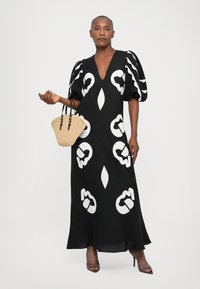 Rochie maxi neagră cu modele abstracte albe, decolteu în V, mâneci puff scurte și tiv evazat. Modelul ține în mână o geantă din paie împletită.