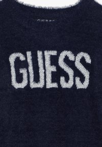 Marineblauer Pullover mit flauschiger Textur und rundem Ausschnitt, der ein auffälliges, weißes, gestricktes "GUESS"-Logo quer über der Brust zeigt.