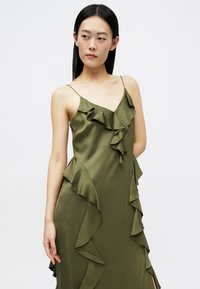 Mulher vestindo um vestido de cetim verde-oliva com alças finas e volantes em cascata, olhando para o lado contra um fundo branco liso.