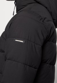 Mustalla puhvitakkilla, jossa on tikkikuvio, varustettuna brändipatchilla hihassa, korkealla kauluksella ja sileällä pinnalla.