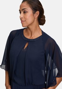 Marinefarbene Bluse mit transparenten Umhangärmel, ausgestattet mit einem runden Ausschnitt, der mit Strasssteinen verziert ist, und einem Schlitzdetail an der Vorderseite.