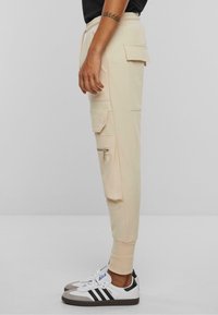 Beige cargo bukser med sidelommer og lynlåse, nedtrappede ben, elastisk talje, sammen med sorte og hvide sneakers. Glat stoftekstur.
