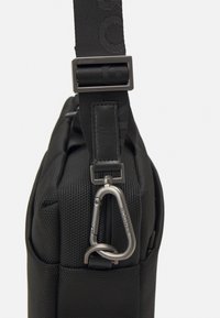 Sort stof skuldertas med struktureret overflade, forsynet med en metalkarabiner og mærket rem, rektangulær form, robust design.