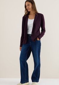 Fialový blazer vyrobený z elastického materiálu, kombinovaný s džíny s vysokým pasem, s volným střihem a bočními kapsami. Neutrální pozadí.