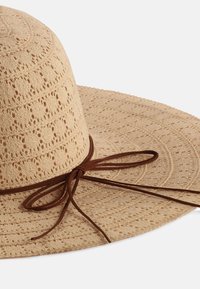 Beżowy kapelusz słomkowy z szerokim rondem, o teksturowanym wzorze i brązową wstążką na dolnej krawędzi. Wykonany z oddychającego, lekkiego materiału.