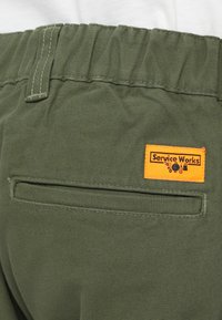 Szorty z zielonej oliwkowej tkaniny z elastycznym pasem, z tylną kieszenią oraz pomarańczową metką "Service Works" z czarną grafiką.