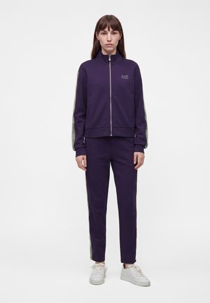 Femme portant un survêtement zippé violet foncé avec des bandes claires sur les manches et le pantalon, associé à des baskets blanches, debout devant un fond uni.