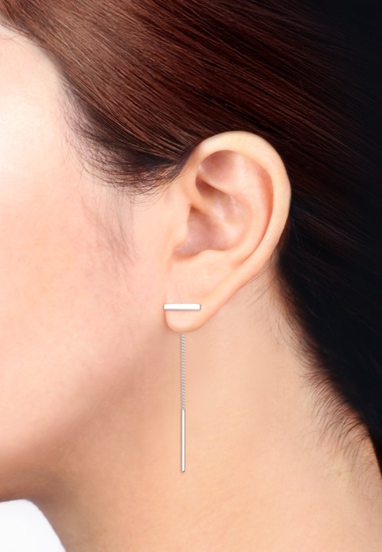 Gros plan sur l'oreille d'une femme aux cheveux lisses châtain roux ramenés derrière, portant une boucle d'oreille pendentif minimaliste en argent avec barre et chaîne.