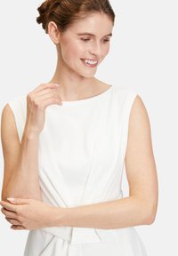 Valkoinen mekko, jossa on veneolkaimet, laskostusvyötäröllä ja istuva yläosa. Sujuva rakenne ja elegantti siluetti.
