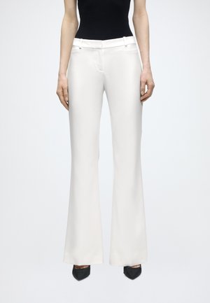 Žena nosiace biele nohavice so širokými nohavicami a vysokým pásom, v kombinácii s čiernymi lodičkami so špicatou špičkou a čiernym tielkom bez rukávov.