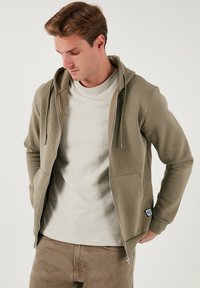 Sudadera verde oliva con cremallera, con un bolsillo delantero, capucha con cordón y una camiseta gris claro debajo. Tejido suave y corte relajado.